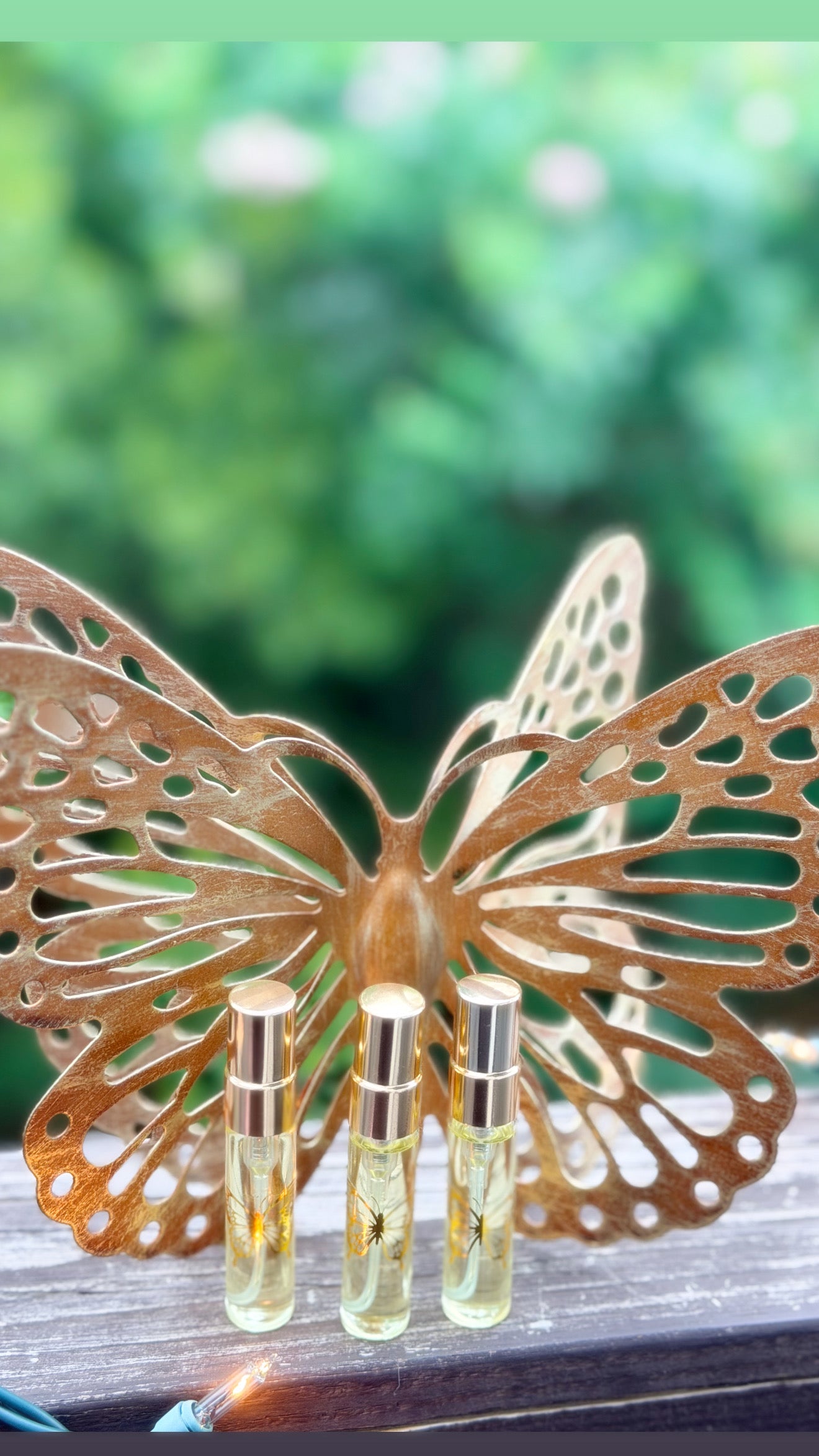 Three small glass perfume or oil vials with silver caps are displayed in front of a decorative butterfly sculpture on a wooden surface, with a blurred green outdoor background.