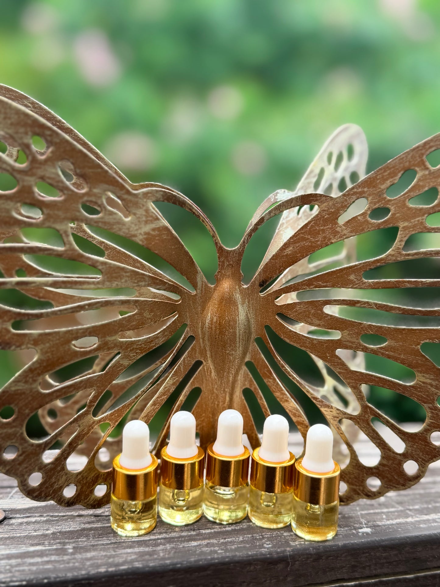 Five small glass dropper bottles filled with golden liquid are lined up in front of a large decorative bronze butterfly sculpture on a wooden surface, with a blurred green outdoor background.
