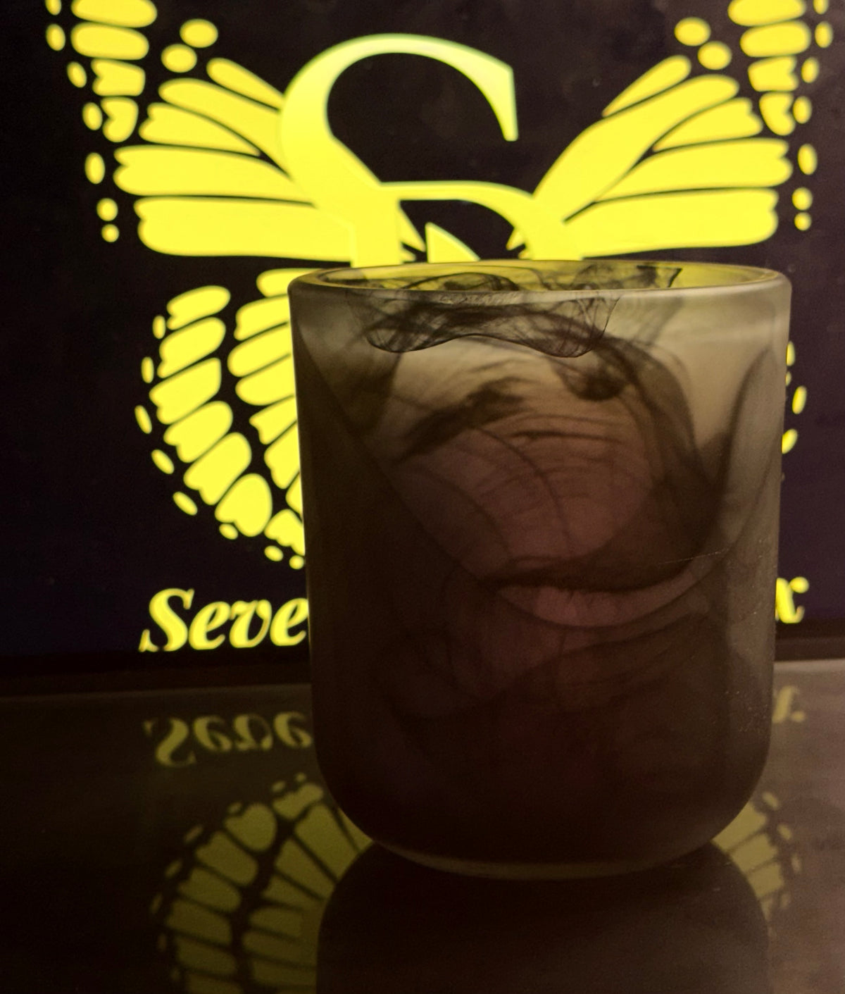 A frosted glass candle holder with a black and gray butterfly wing design, shown on a reflective surface with a glowing yellow butterfly backdrop.
