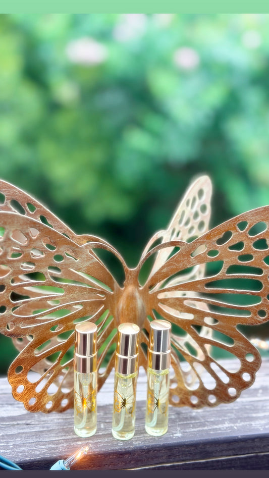 Three small glass perfume or oil vials with silver caps are displayed in front of a decorative butterfly sculpture on a wooden surface, with a blurred green outdoor background.
